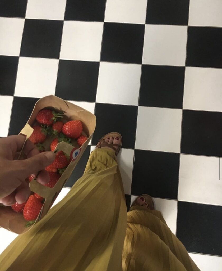 Geste du quotidien capté sur un sol graphique : robe fluide moutarde, panier de fraises et damier noir et blanc. Symbole d'un alignement entre corps, espace et rythme de vie.